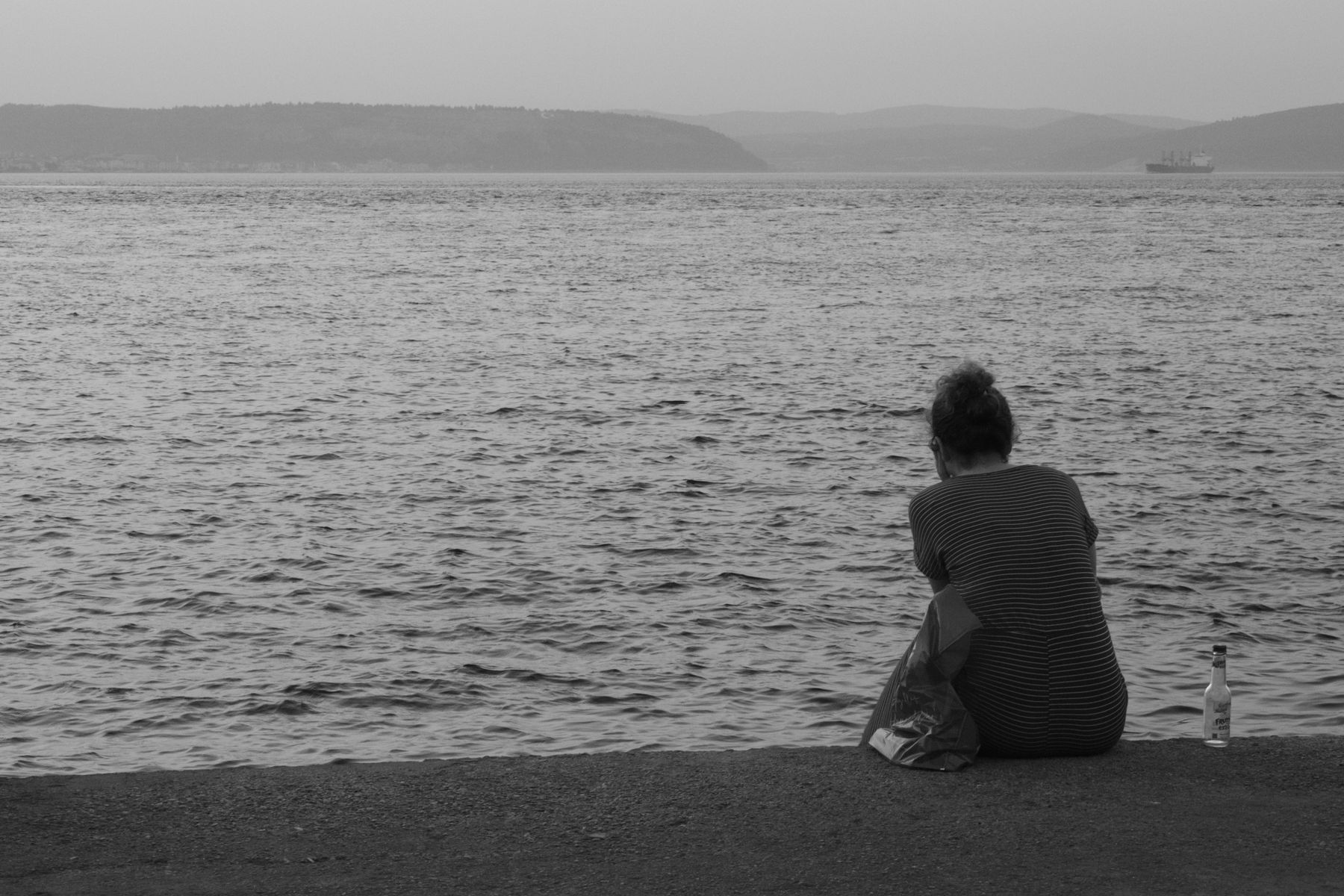 A solitary figure seated on a quiet pier looking out over calm water toward distant hills