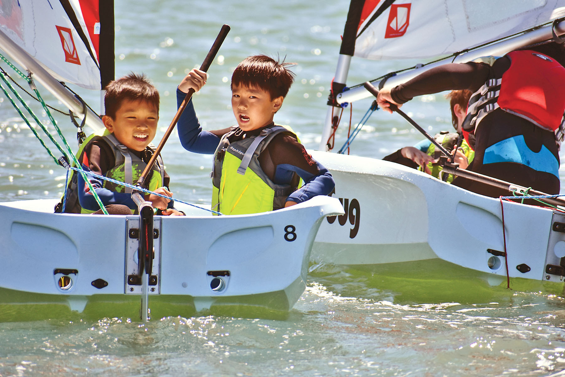 Two young sailors smiling in life vests aboard small dinghies on bright open water